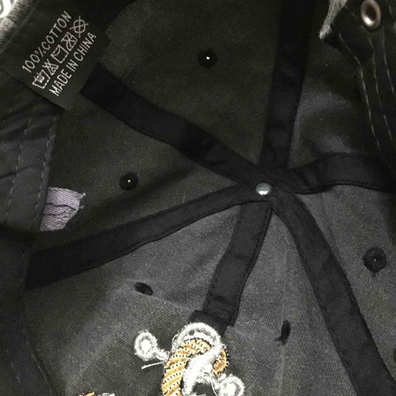 Close-up of the interior of a black baseball cap, showing the adjustable black fabric straps, metal snap closure, ventilation eyelets, and a small embroidered patch near the lower front.