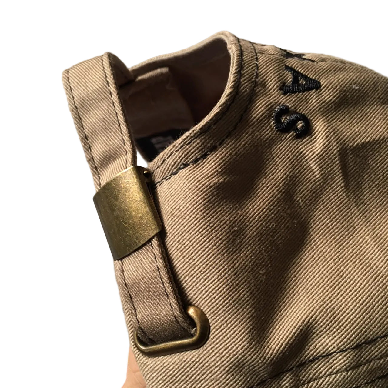 Close-up of a beige canvas hat with a brass-colored metal strap and a black embroidered logo.